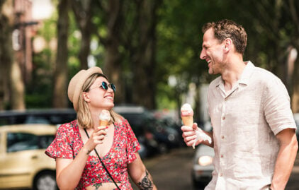 Eine junge Frau und ein junger Mann essen ein Eis und scheinen sich sehr fröhlich zu unterhalten. Sie trägt einen Sonnenhut und ein rotes Sommerkleid. Der Mann trägt ein beiges Hemd.